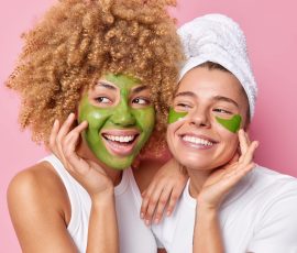 Photo of happy two women touch face enjoy softness of skin undergo beauty procedures smile broadly apply mask and patches have glad expressions isolated over pink background. Facial treatment